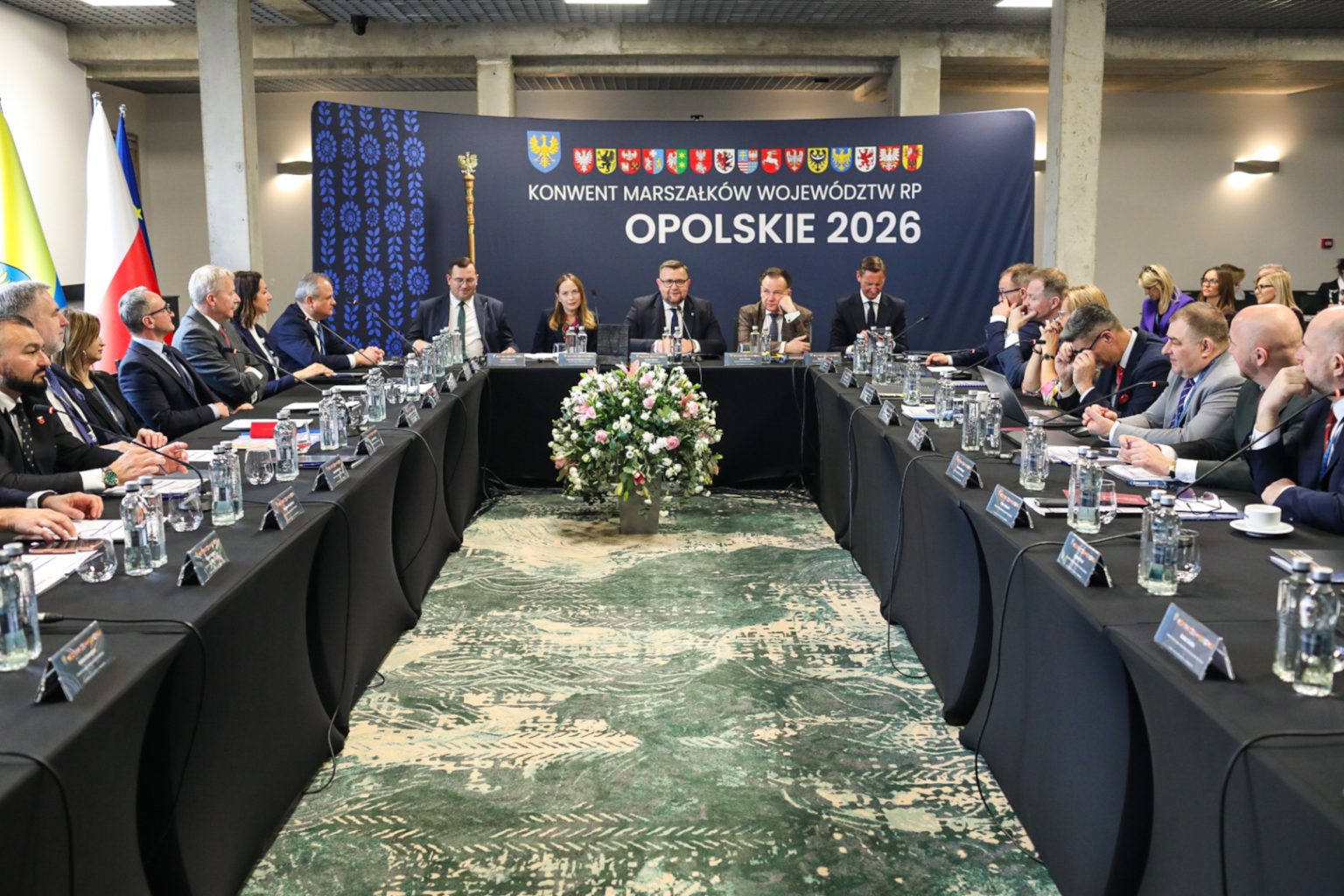 Zdjęcie dużej sali. Po dwóch stronach naprzeciwko siebie ustawione są stoliki z krzesłami, przy których siedzą Marszałkowie Polskich Województw. Centralnie widoczny jest stół prezydialny, przy którym siedzi 5 osób, jedna kobieta i czterech mężczyzn. Z tyłu widoczna jest ścianka z napisem: „Konwent Marszałków Województw RP, Opolskie 2026”.