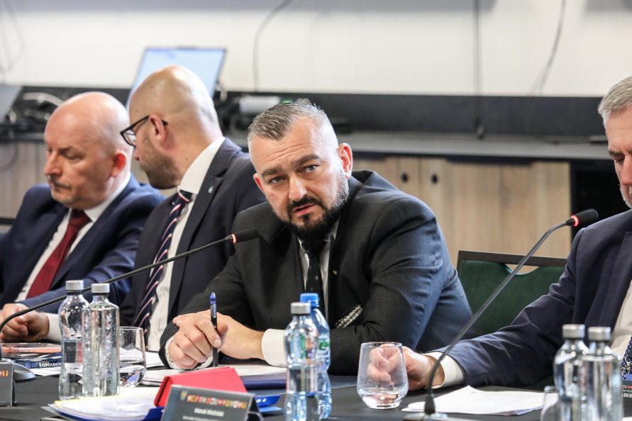 Centralnie na zdjęciu widoczny jest, ubrany w garnitur i białą koszulę, mężczyzna, który siedzi przy stole. Jest to Jarosław Kwasek, Członek Zarządu Województwa Lubelskiego. Po jego obu stronach siedzą inni mężczyźni.