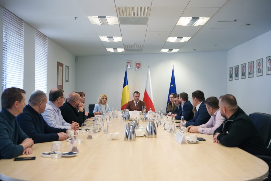 Spotkanie delegacji z Członkiem Zarządu Województwa Lubelskiego Marcinem Szewczakiem. Uczestnicy spotkania siedzą przy stole konferencyjnym, za nimi widoczne są flagi Rumunii, Polski i Unii Europejskiej.