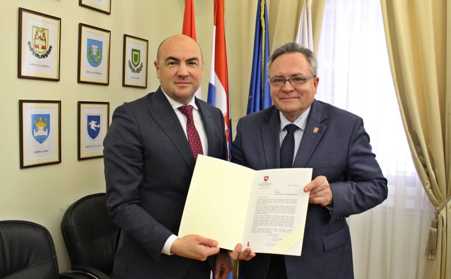 Pan Blaž Pezo Żupan Dubrownicko-neretwiańskiej Żupanii oraz Pan Zbigniew Wojciechowski Przewodniczący Komisji Rozwoju Regionalnego i Współpracy Zagranicznej trzymają razem dokument przed flagami.