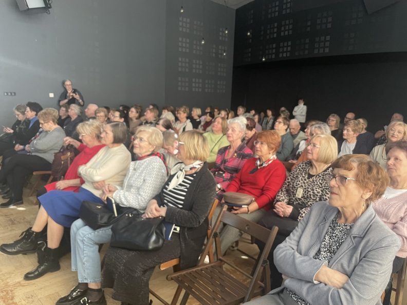 Seniorzy siedzą na sali na krzesłach, kilkadziesiąt osób