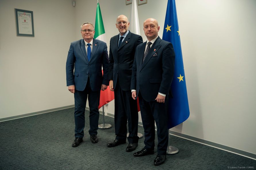 Trzech mężczyzn w garniturach stoi obok siebie pozując do zdjęcia. Za nimi widoczne flagi Włoch, Polski oraz Unii Europejskiej.