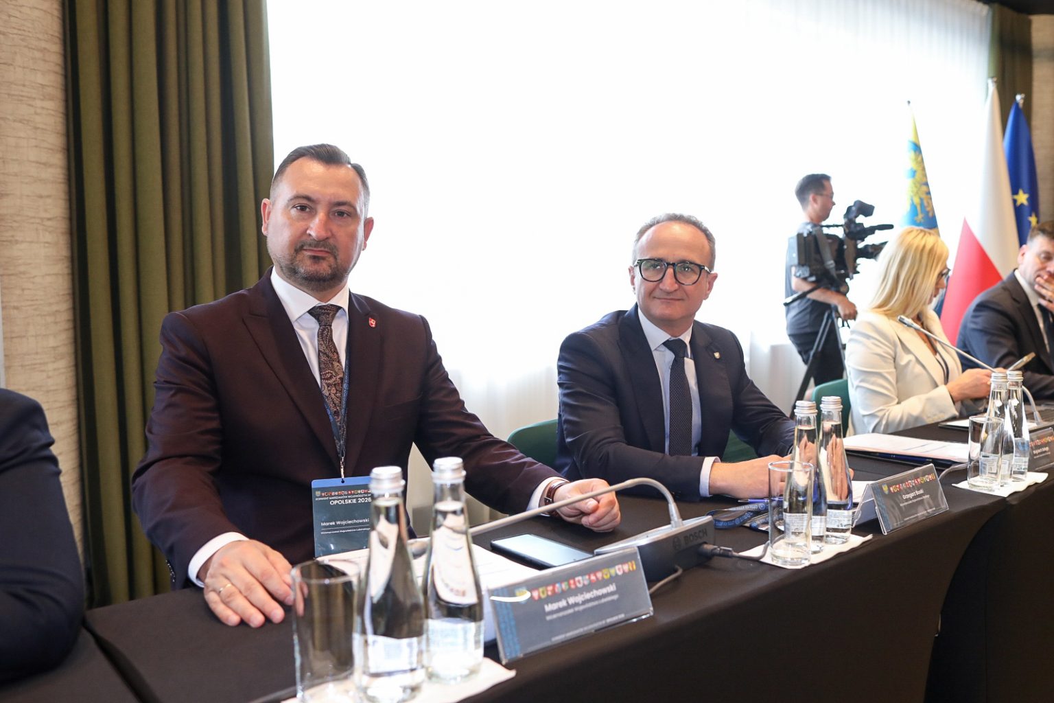 Centralnie na zdjęciu widoczny jest, ubrany w garnitur i białą koszulę, mężczyzna, który siedzi przy stole nakrytym czarnym naciągiem. Jest to Marek Wojciechowski, Wicemarszałek Województwa Lubelskiego. Po prawej stronie siedzi inny mężczyzna, również ubrany w ciemny garnitur i jasną koszulę.