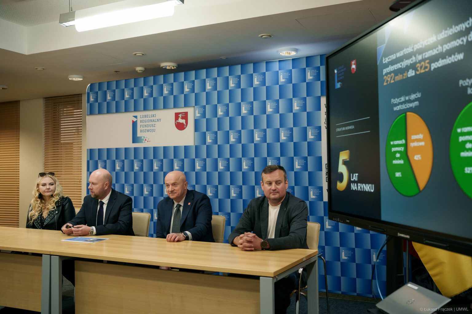 Członkowie konferencji siedzący przy długim stole. Za nimi ozdobna ścianka z logotypami organizatorów.