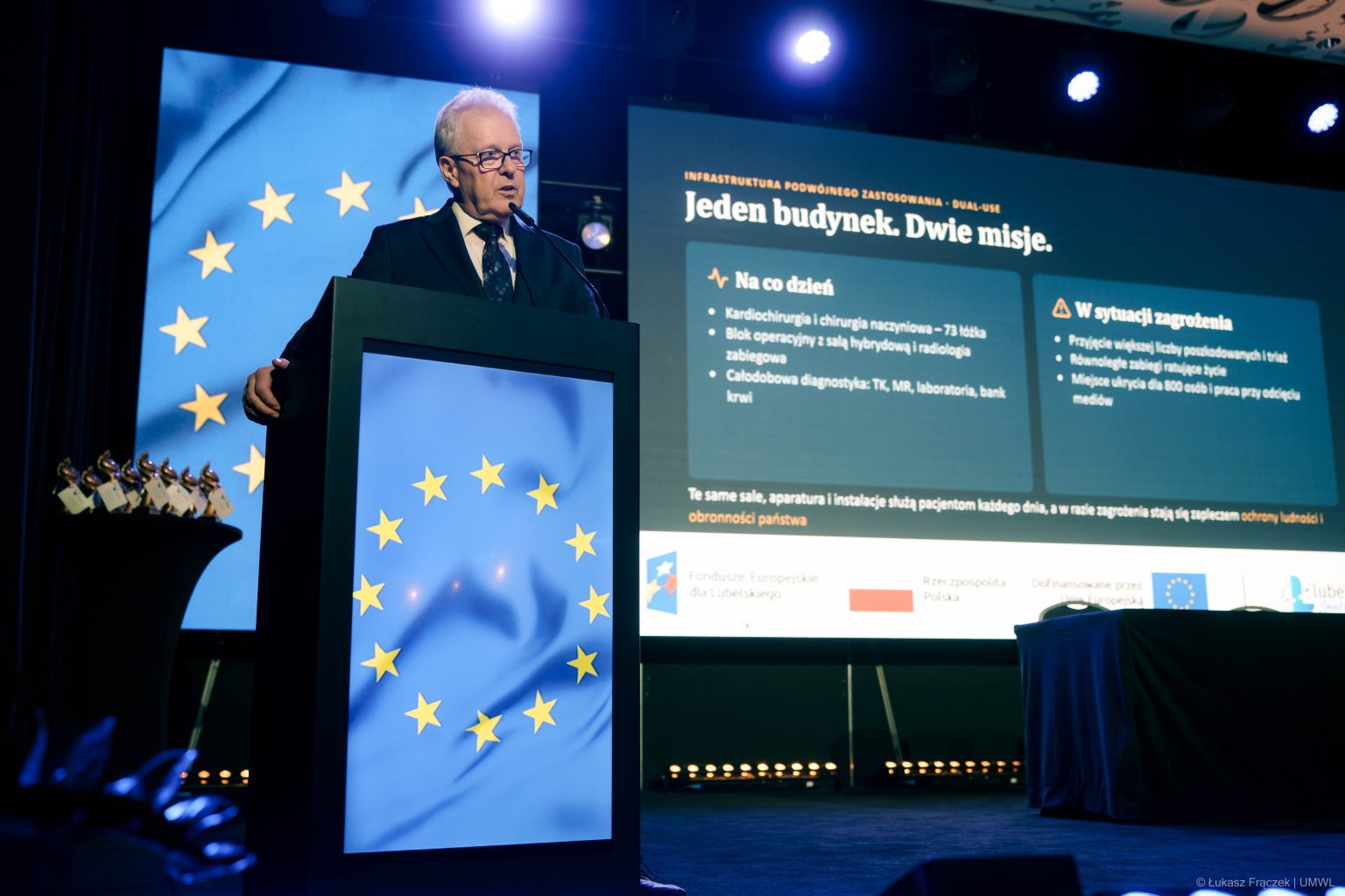 Mężczyzna przemawiający z mównicy. W tle wyświetlona jest prezentacja i wszędzie naokoło widoczna jest flaga Unii Europejskiej.